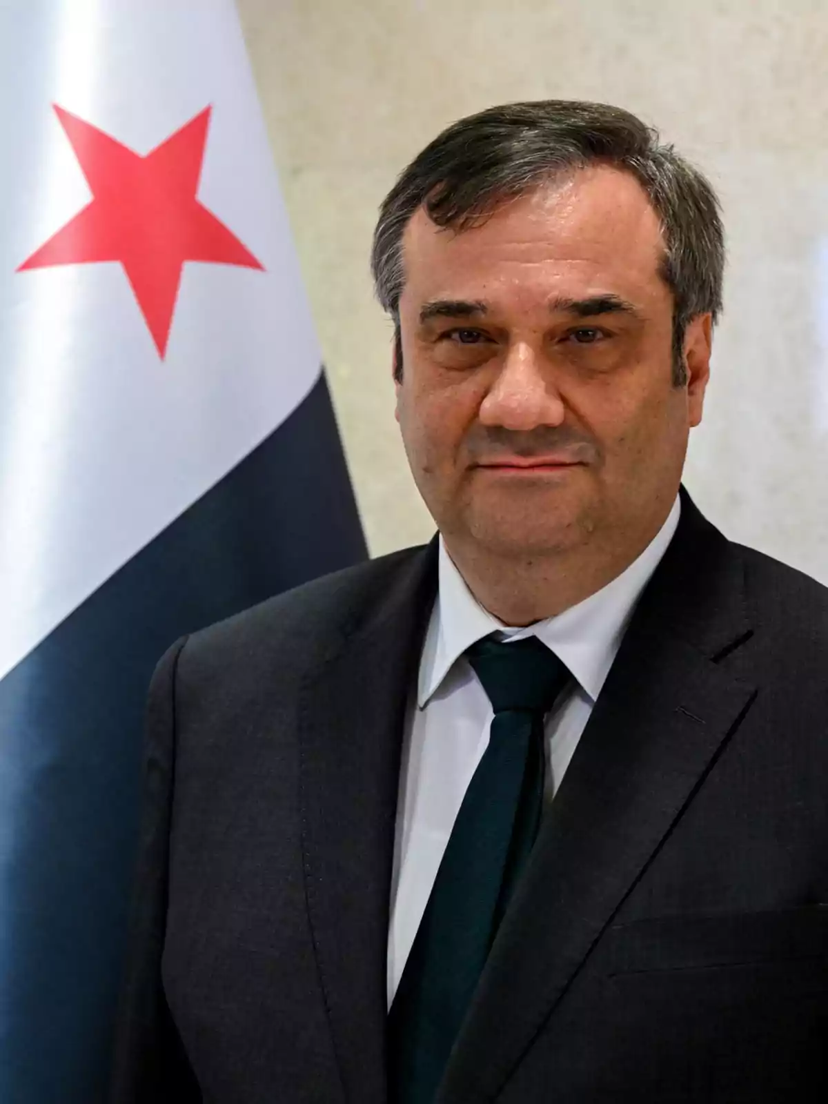 Hombre de mediana edad con cabello corto y canoso vistiendo traje oscuro y corbata, posando frente a una bandera blanca con una estrella roja Hombre de mediana edad con cabello corto y canoso vistiendo traje oscuro y corbata, posando frente a una bandera blanca con una estrella roja