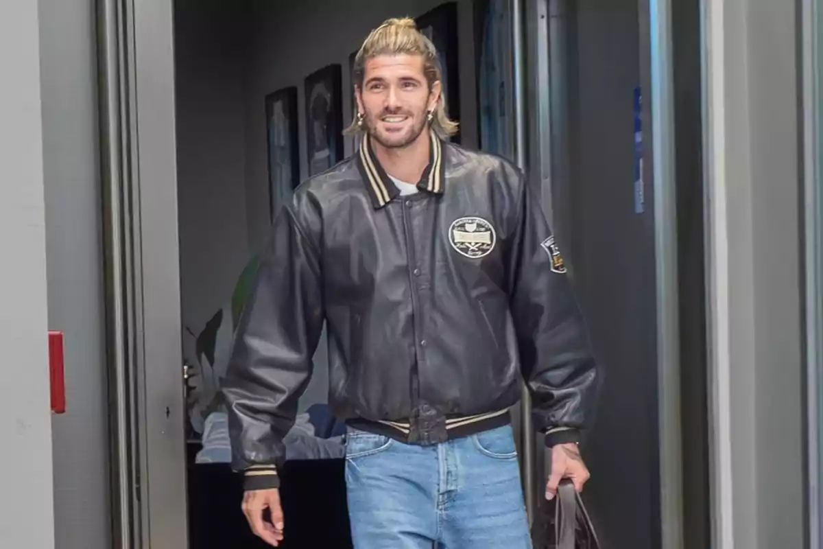 Man in a black leather jacket and light jeans walking down an indoor hallway while smiling