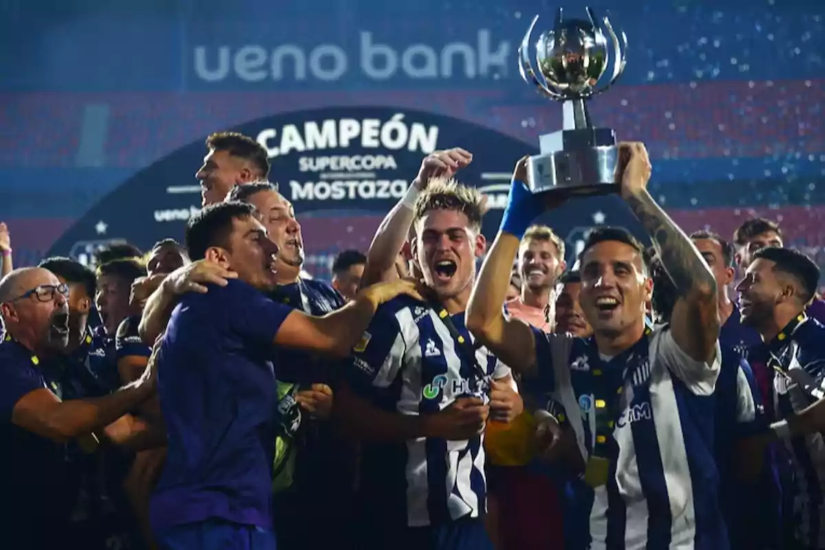 Un equipo de fútbol celebra con un trofeo en alto mientras los jugadores sonríen y se abrazan en un estadio.