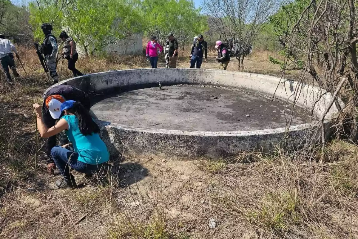 Un grupo de personas, algunas con equipo táctico, exploran un área al aire libre con vegetación seca y un tanque circular de concreto.