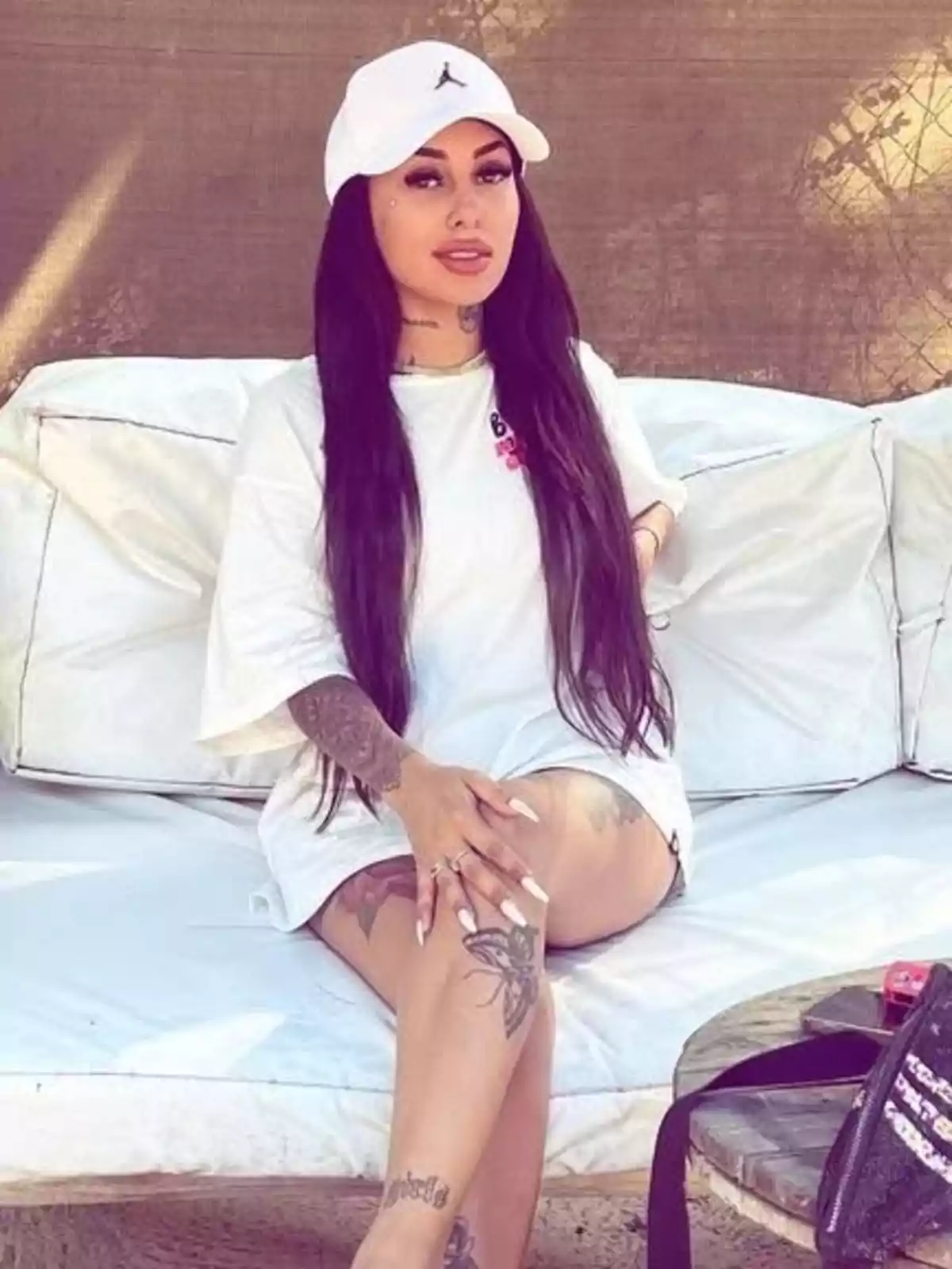 Woman with long dark hair sitting on a white sofa outdoors, wearing a white cap and white t-jersey, with several visible tattoos on her arms and legs. Woman with long dark hair sitting on a white sofa outdoors, wearing a white cap and white t-jersey, with several visible tattoos on her arms and legs.