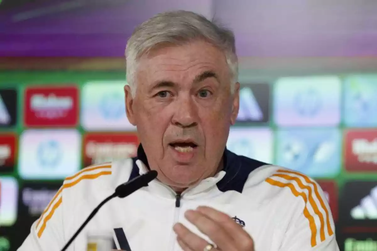 A gray-haired man in a white sports jacket speaks at a press conference with a microphone in front of him and a background of colorful logos. A gray-haired man in a white sports jacket speaks at a press conference with a microphone in front of him and a background of colorful logos.