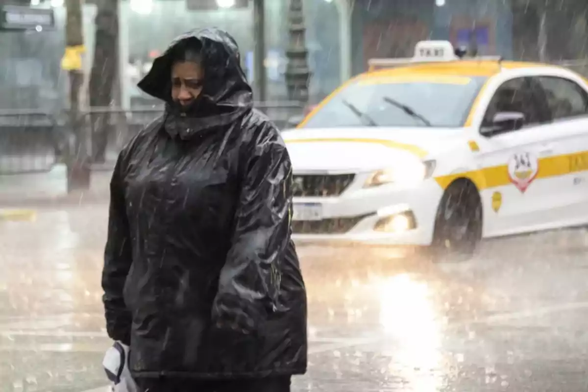 A person in a black raincoat walks in the heavy rain while a taxi passes behind.
