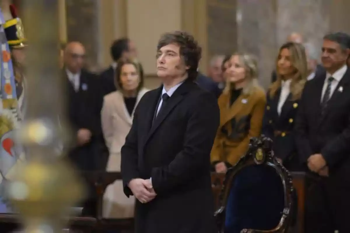 Curly-haired man in a dark suit standing at a formal event with several people in the background inside an elegant building Curly-haired man in a dark suit standing at a formal event with several people in the background inside an elegant building