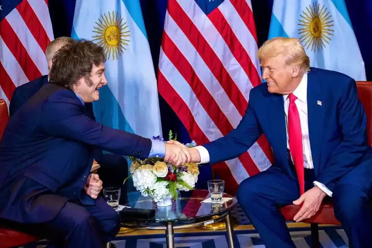 Dos personas dándose la mano en un entorno formal con banderas de Argentina y Estados Unidos de fondo.