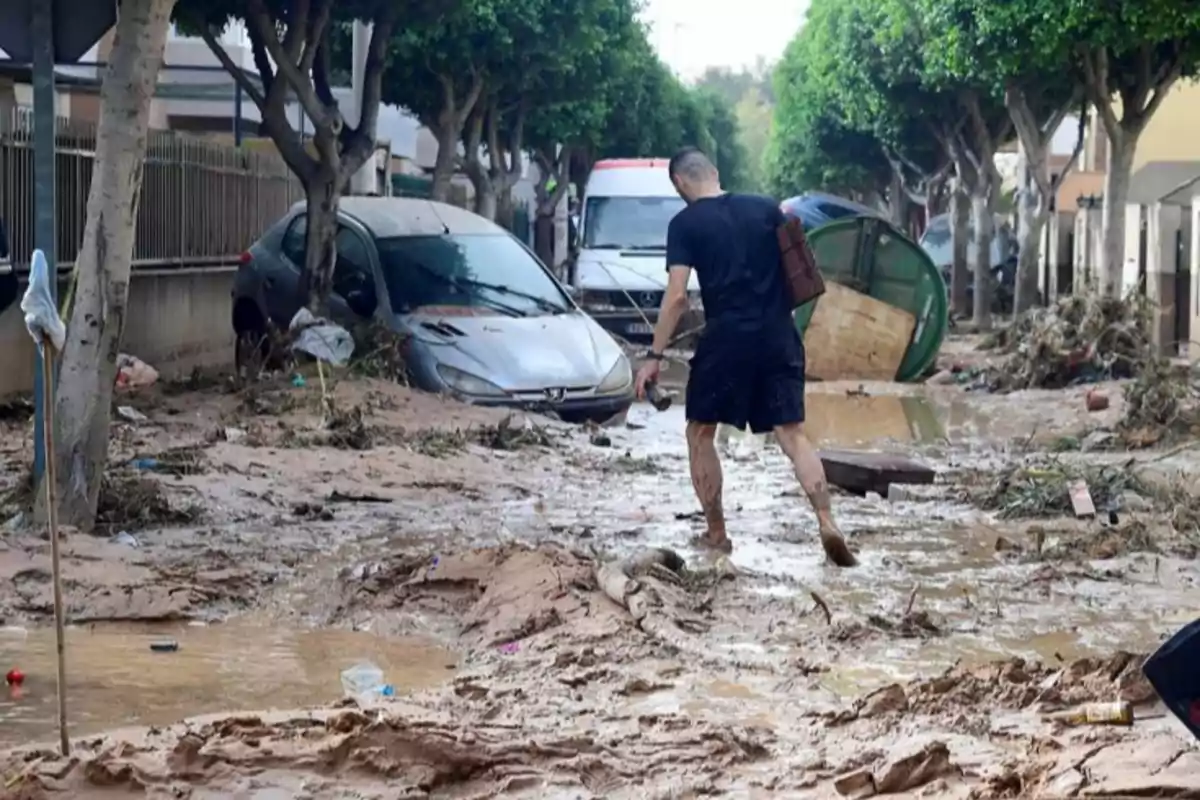 Un hombre camina por una calle inundada y llena de barro con autos y escombros alrededor después de una tormenta.