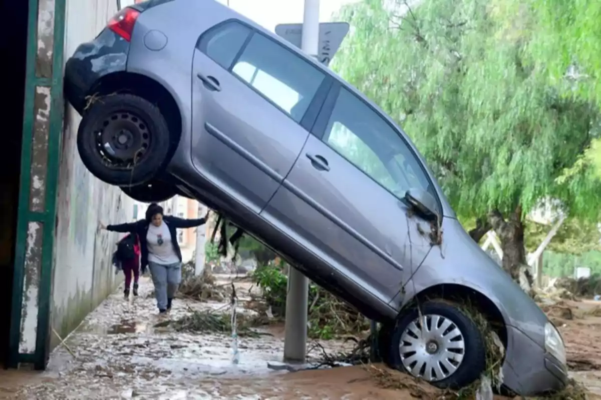 Un coche gris está inclinado contra una pared después de una inundación mientras dos personas caminan por el agua y el lodo.