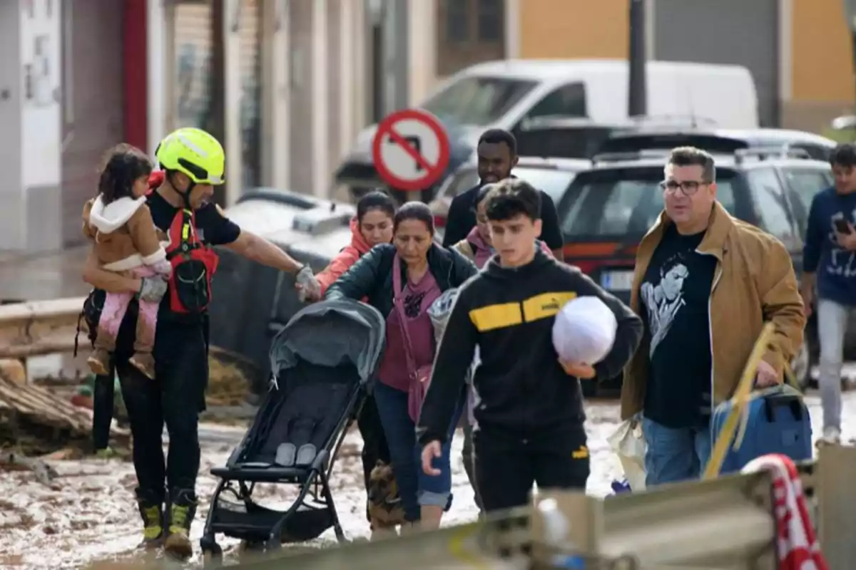 Un grupo de personas es asistido por un rescatista mientras caminan por una calle inundada llevando pertenencias y un cochecito de bebé.