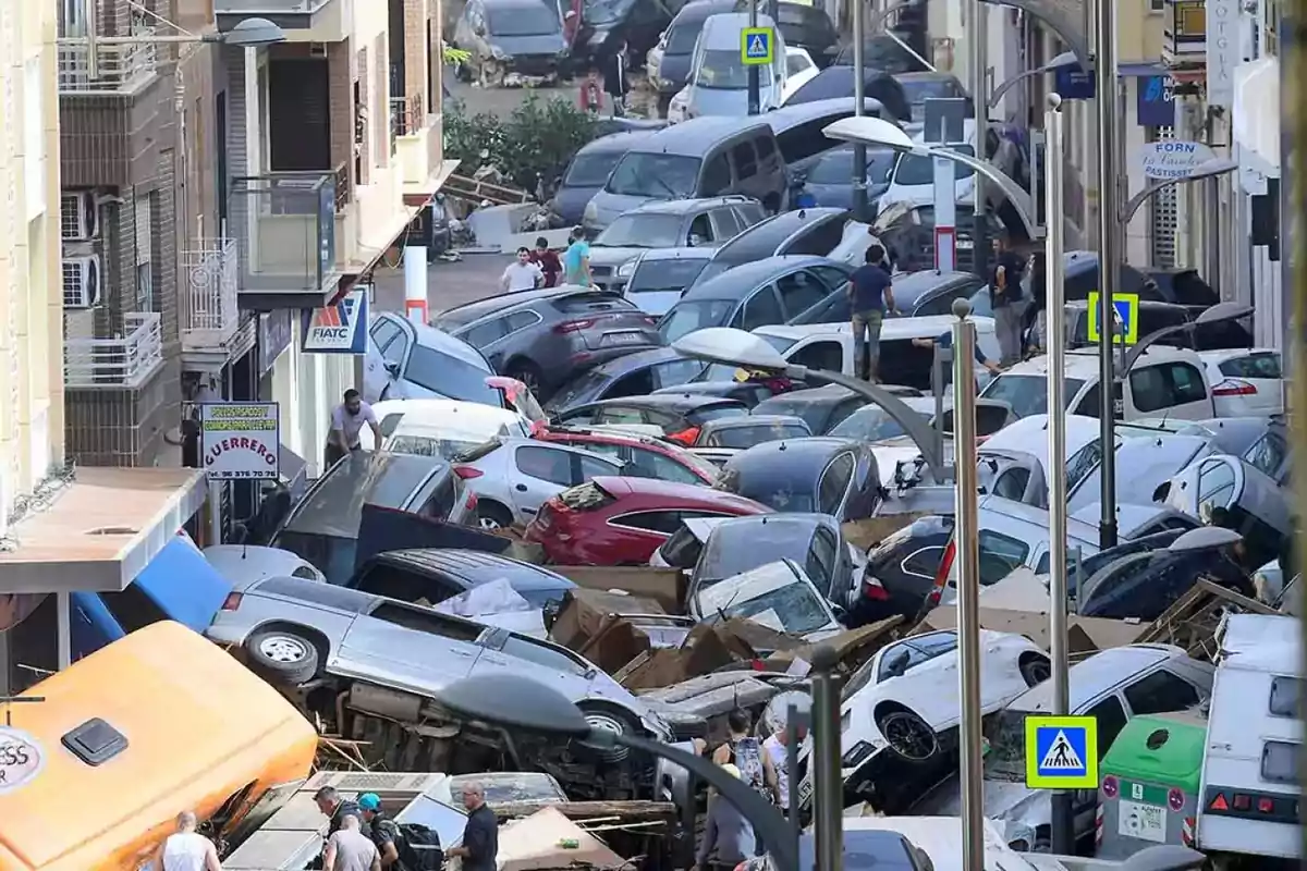 Calle llena de coches apilados y dañados tras una inundación, con personas caminando entre ellos.