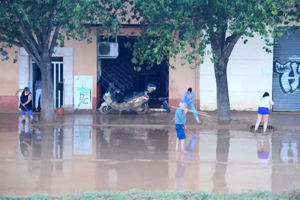 Personas limpiando una calle inundada con escobas y palas frente a un edificio con árboles y una motocicleta estacionada.