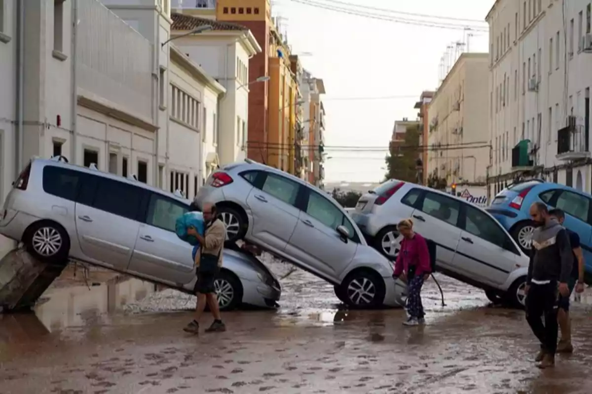 Coches apilados en una calle inundada mientras personas caminan por el lugar.