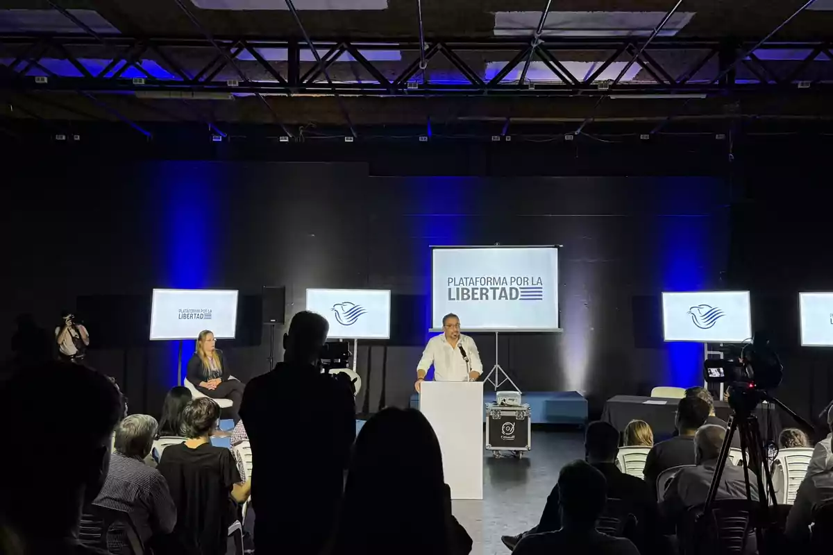 A person speaking at a podium during an event with screens displaying the text A person speaking at a podium during an event with screens displaying the text
