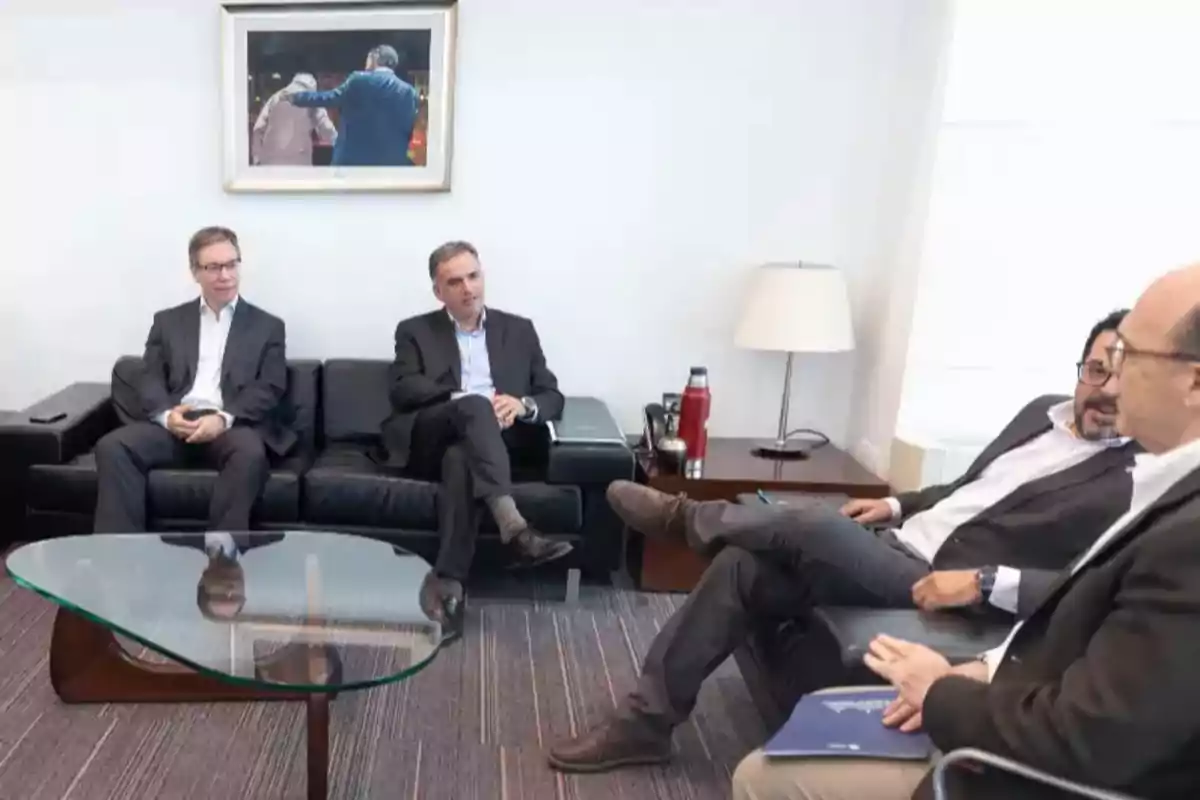 Four people in formal suits are sitting on black sofas in a modern meeting room with a glass table and a lamp, while conversing. Four people in formal suits are sitting on black sofas in a modern meeting room with a glass table and a lamp, while conversing.