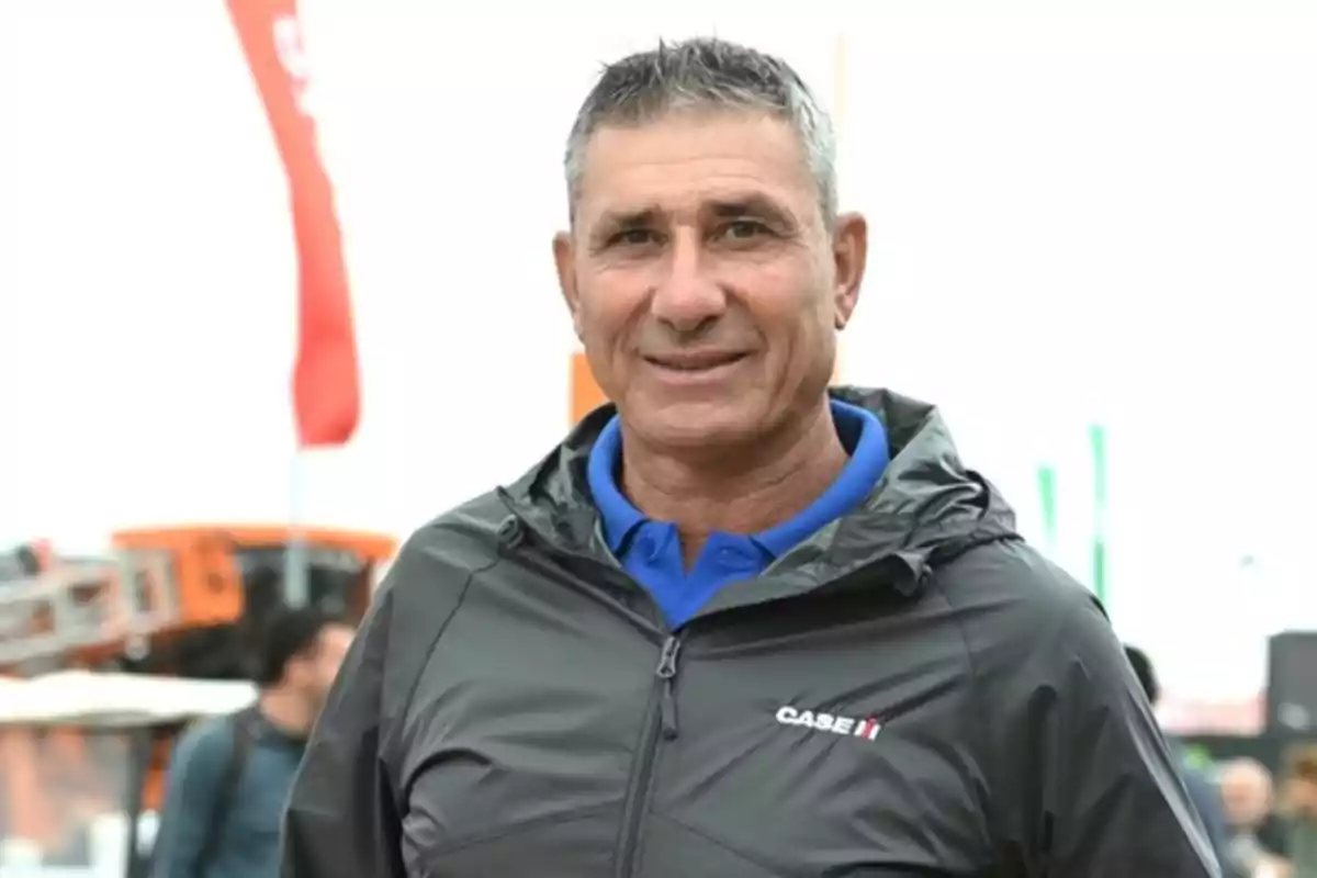 A man in a black jacket and blue jersey smiling outdoors with a blurry background.