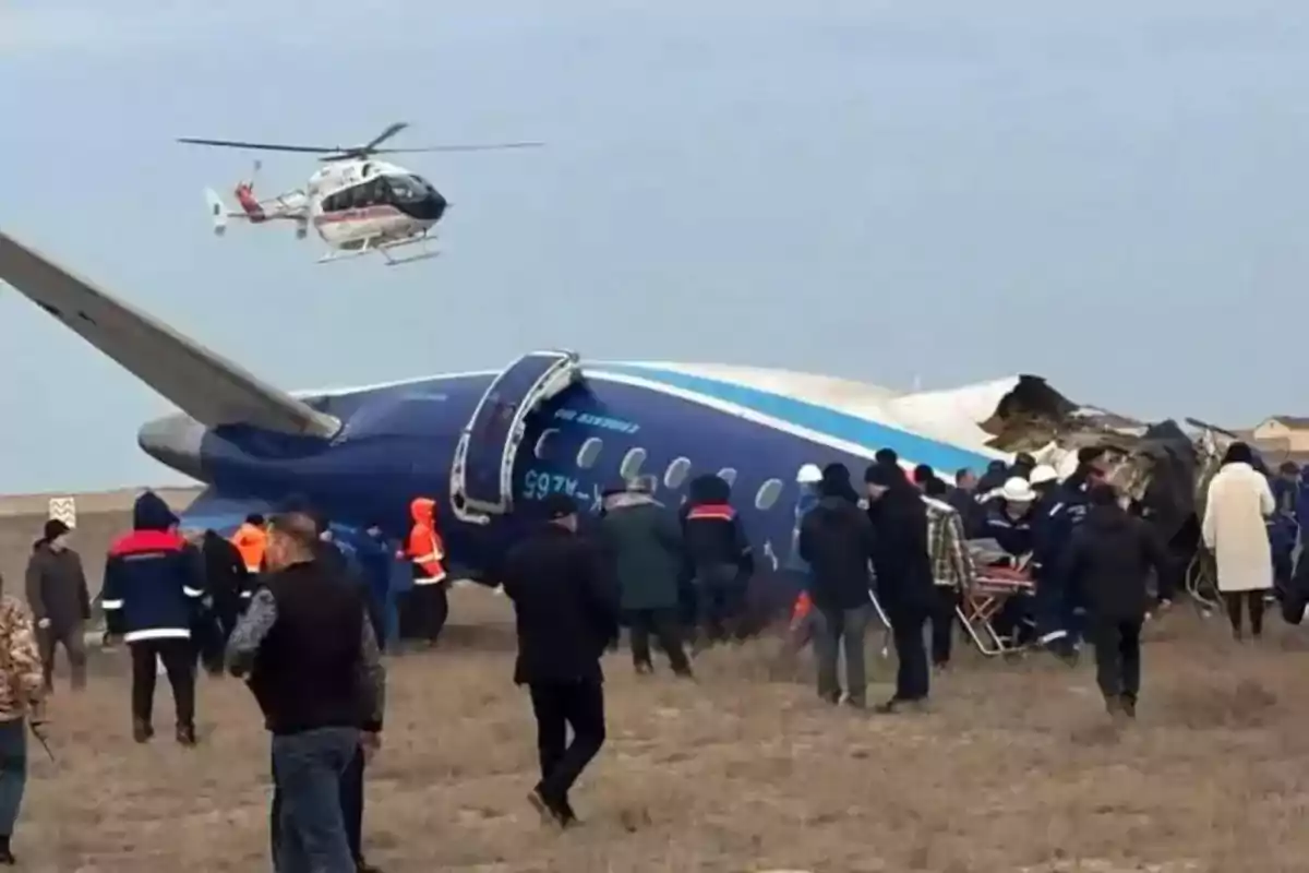 Un grupo de personas y rescatistas rodea un avión accidentado en un campo mientras un helicóptero sobrevuela la escena.