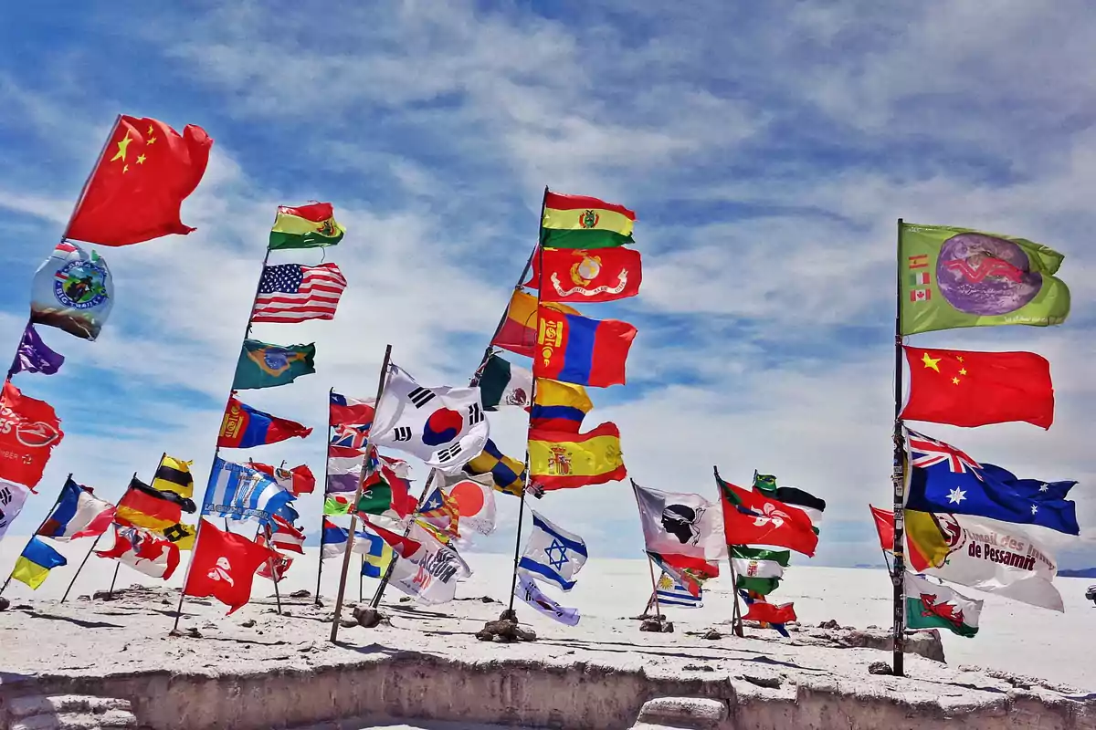 Un grupo de banderas de diferentes países ondea al viento en un paisaje desértico bajo un cielo azul.