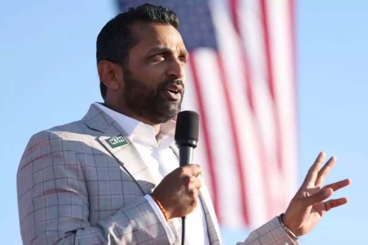 A man speaking with a microphone in hand, wearing a plaid suit, with a flag in the background. A man speaking with a microphone in hand, wearing a plaid suit, with a flag in the background.