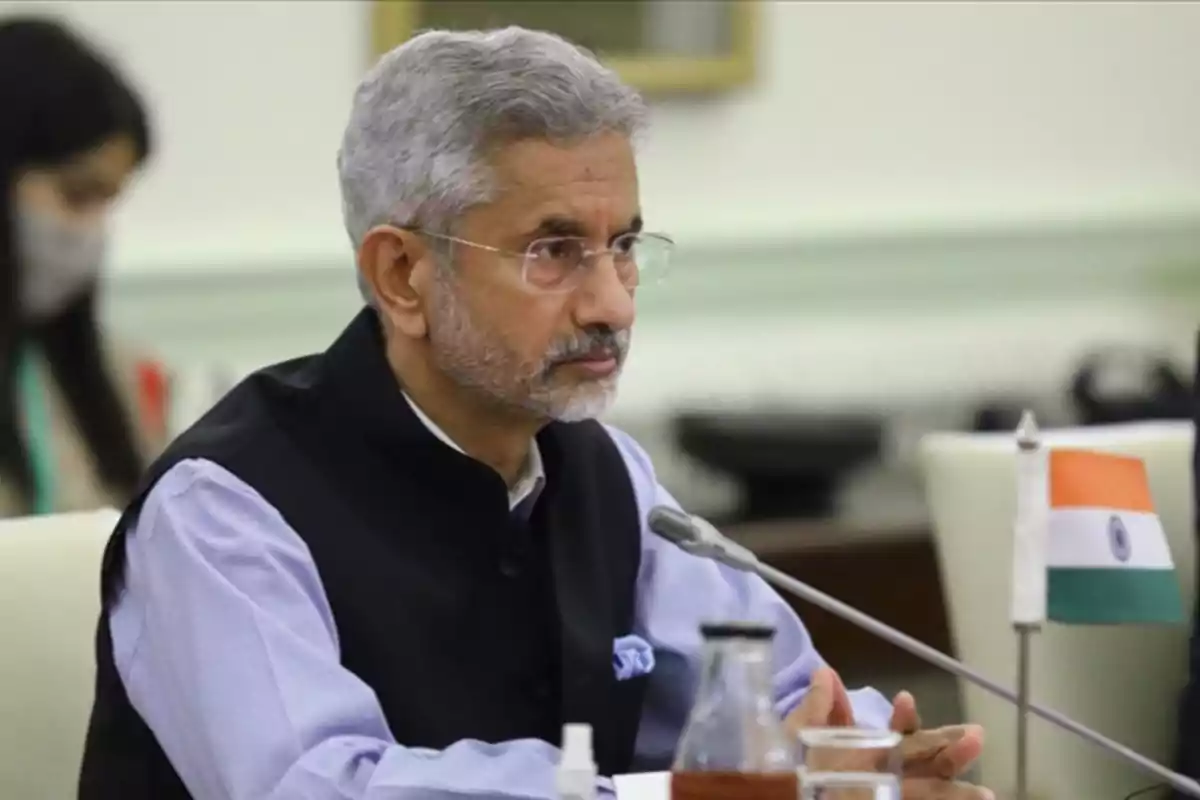 A gray-haired man with a beard is sitting at a table with a microphone in front of him and a small Indian flag beside him. A gray-haired man with a beard is sitting at a table with a microphone in front of him and a small Indian flag beside him.