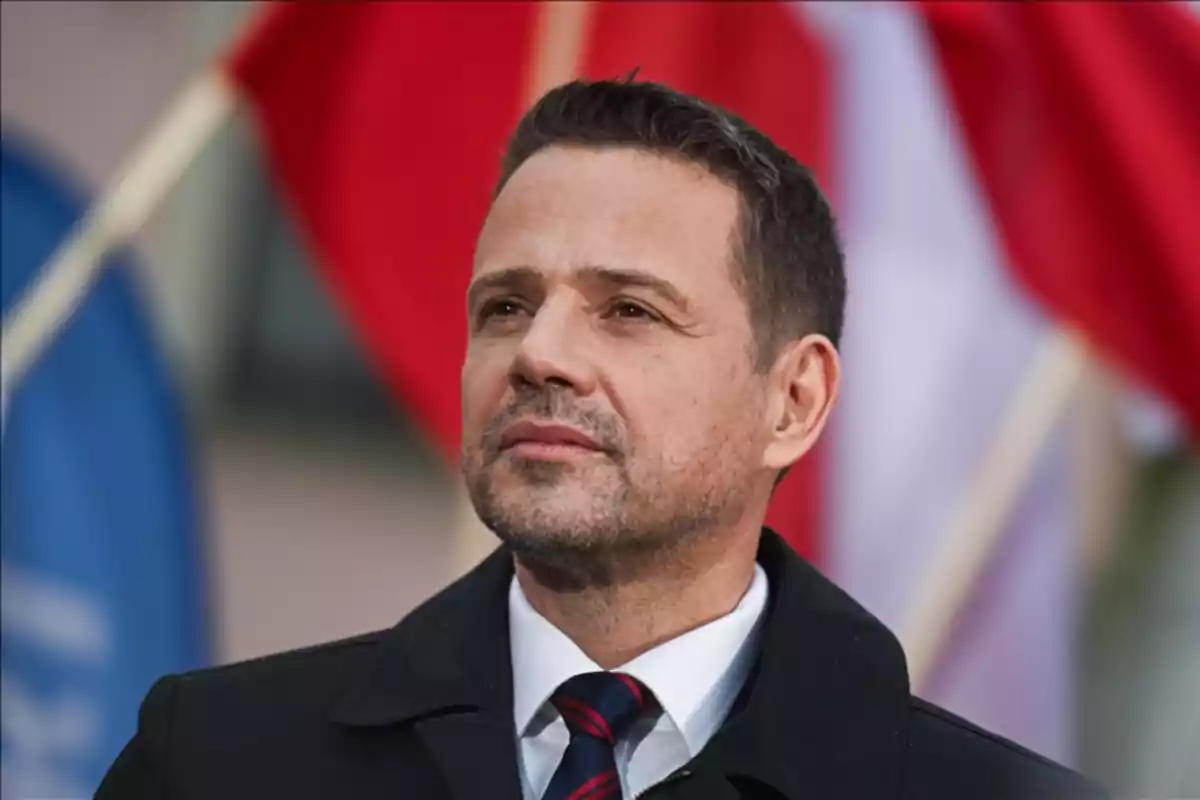 Hombre de cabello corto y barba incipiente con traje formal y corbata, de pie frente a banderas desenfocadas en el fondo Hombre de cabello corto y barba incipiente con traje formal y corbata, de pie frente a banderas desenfocadas en el fondo