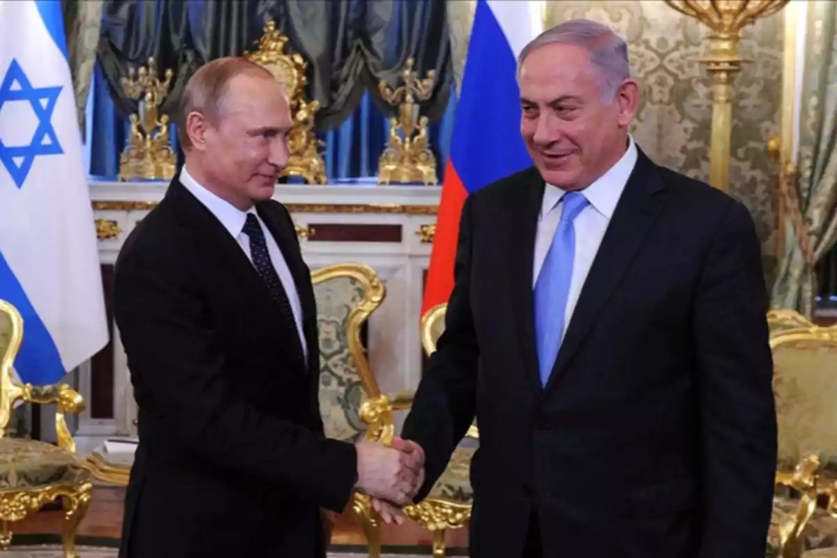 Two men in suits shake hands in an elegant room with Israeli and Russian flags in the background. Two men in suits shake hands in an elegant room with Israeli and Russian flags in the background.