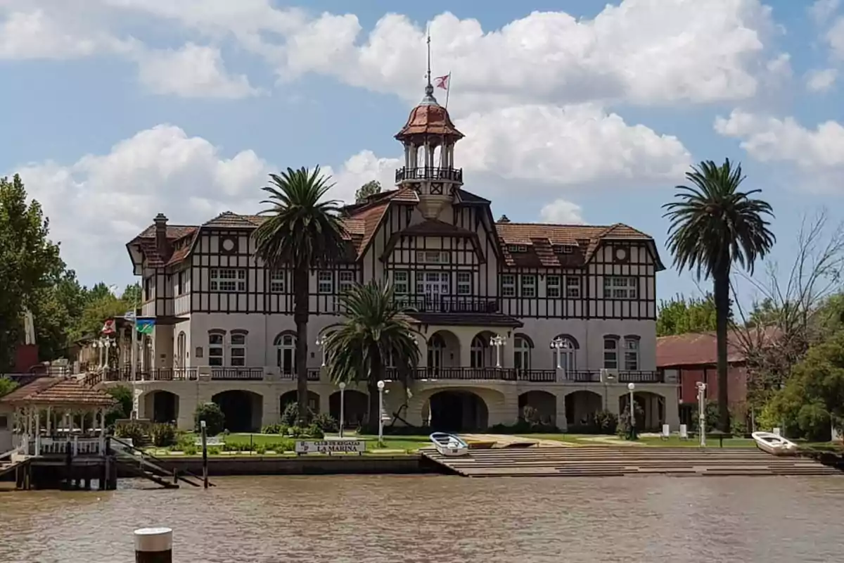 Un edificio histórico de estilo europeo con techo de tejas rojas y una torre central rodeado de palmeras junto a un río bajo un cielo azul con nubes.