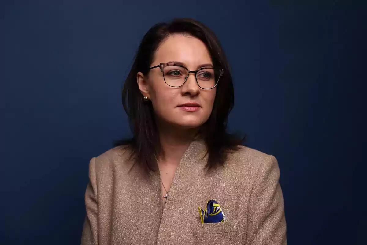 Una persona con gafas y cabello oscuro lleva un saco beige con un pañuelo azul en el bolsillo sobre un fondo azul. Una persona con gafas y cabello oscuro lleva un saco beige con un pañuelo azul en el bolsillo sobre un fondo azul.
