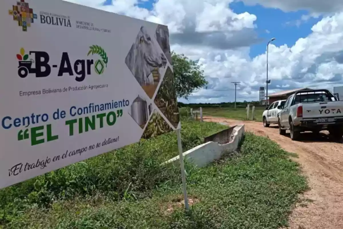 Un cartel de la Empresa Boliviana de Producción Agropecuaria "B-Agro" que indica el "Centro de Confinamiento El Tinto" junto a una carretera de tierra y dos camionetas estacionadas, con un cielo parcialmente nublado al fondo.