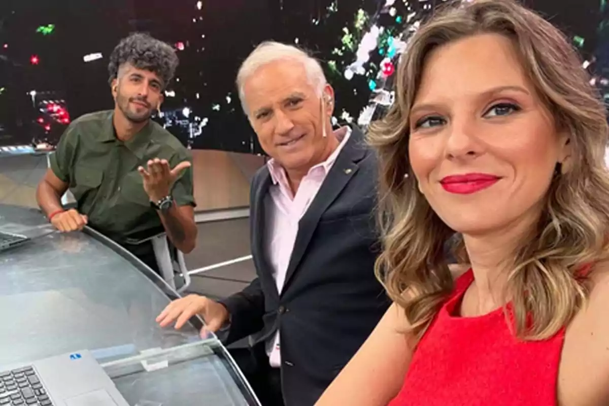 Tres personas posan sonrientes en un estudio de televisión, una mujer lleva un vestido rojo, un hombre mayor viste un traje oscuro y otro hombre más joven lleva una camisa verde. Tres personas posan sonrientes en un estudio de televisión, una mujer lleva un vestido rojo, un hombre mayor viste un traje oscuro y otro hombre más joven lleva una camisa verde.