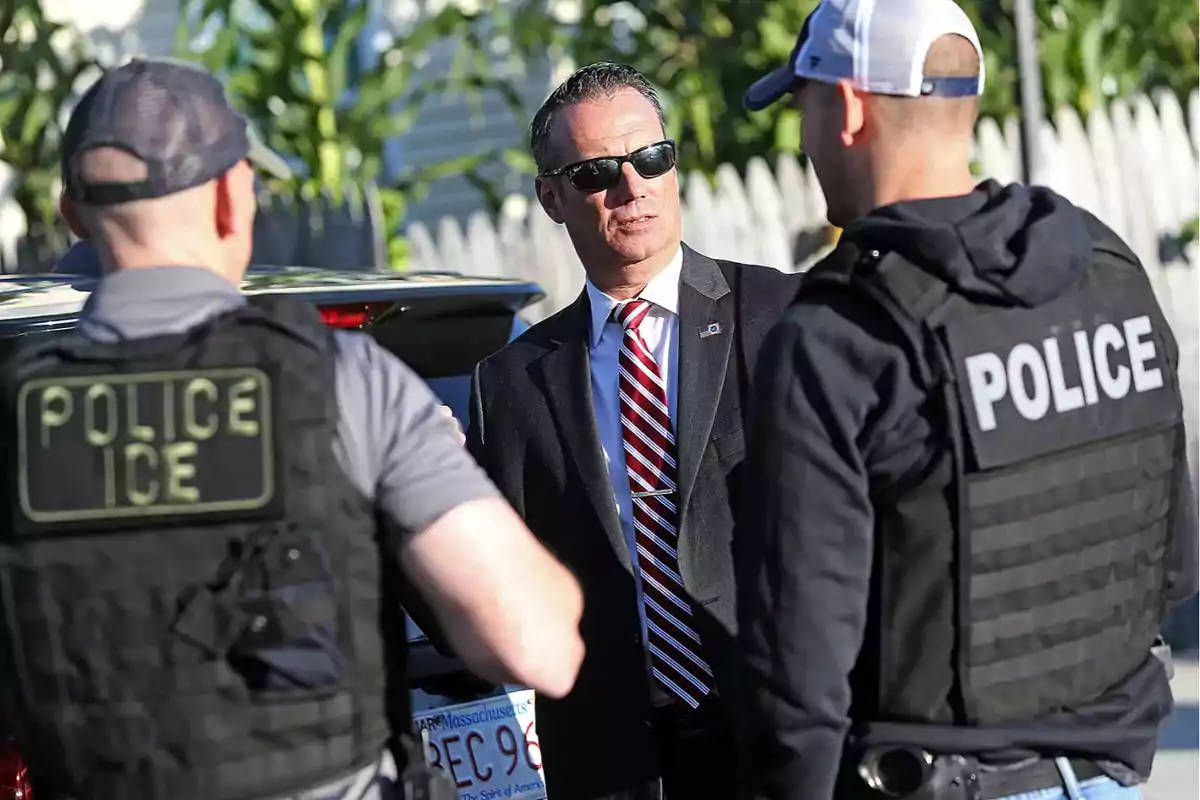 Three police officers, two wearing bulletproof vests and one in a suit with sunglasses, are talking next to a vehicle in a residential area during the day. Three police officers, two wearing bulletproof vests and one in a suit with sunglasses, are talking next to a vehicle in a residential area during the day.