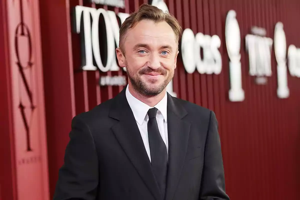 Man in a black suit and tie smiling while posing on a red carpet in front of a backdrop with the Tony Awards logo Man in a black suit and tie smiling while posing on a red carpet in front of a backdrop with the Tony Awards logo