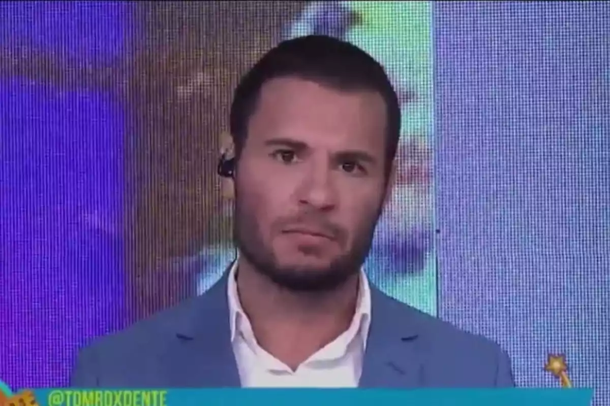 Un hombre con auriculares y chaqueta azul aparece en un programa de televisión con un fondo colorido.