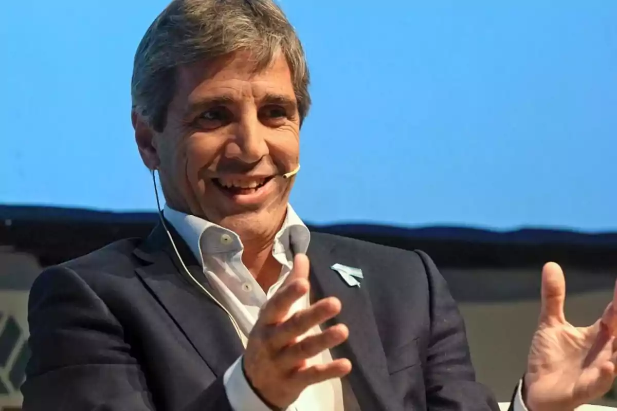 Un hombre con traje oscuro y camisa blanca, usando un micrófono de diadema, gesticula y sonríe mientras habla en un escenario con fondo azul.