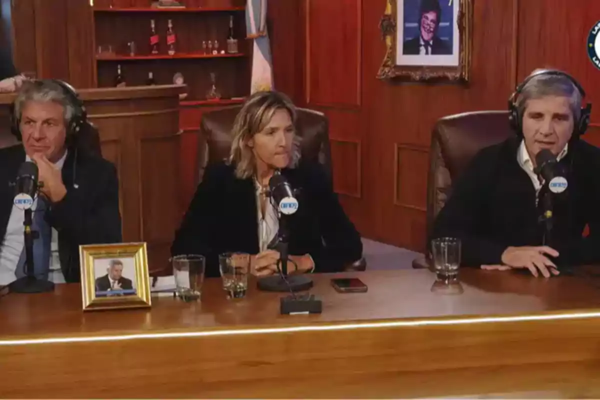 Three people with headphones are sitting at a wooden table with microphones in front of them, in a studio with shelves and an Argentine flag in the background. Three people with headphones are sitting at a wooden table with microphones in front of them, in a studio with shelves and an Argentine flag in the background.