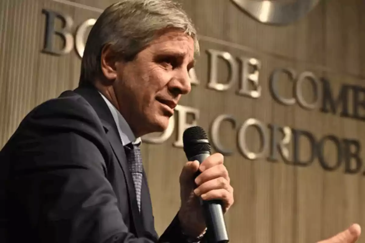 Gray-haired man in a dark suit speaking into a microphone at an institutional event with letters on the wall in the background. Gray-haired man in a dark suit speaking into a microphone at an institutional event with letters on the wall in the background.