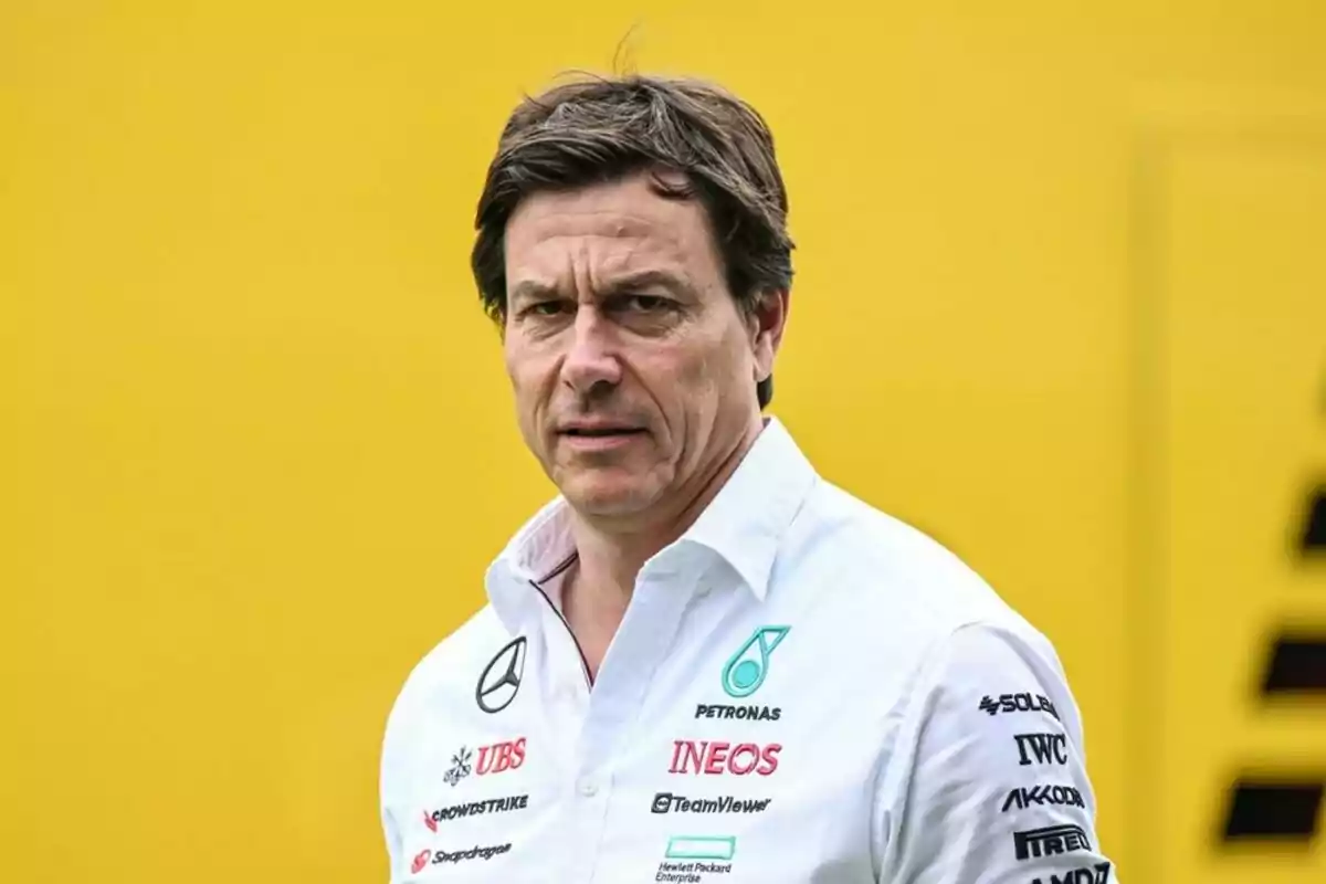 Un hombre con camisa blanca con logotipos de patrocinadores está de pie frente a un fondo amarillo. Un hombre con camisa blanca con logotipos de patrocinadores está de pie frente a un fondo amarillo.