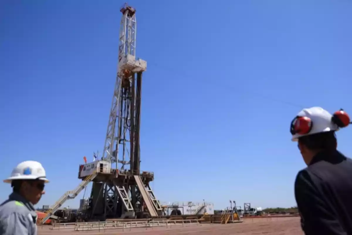 Personas con cascos observan una torre de perforación petrolera en un campo bajo un cielo despejado.