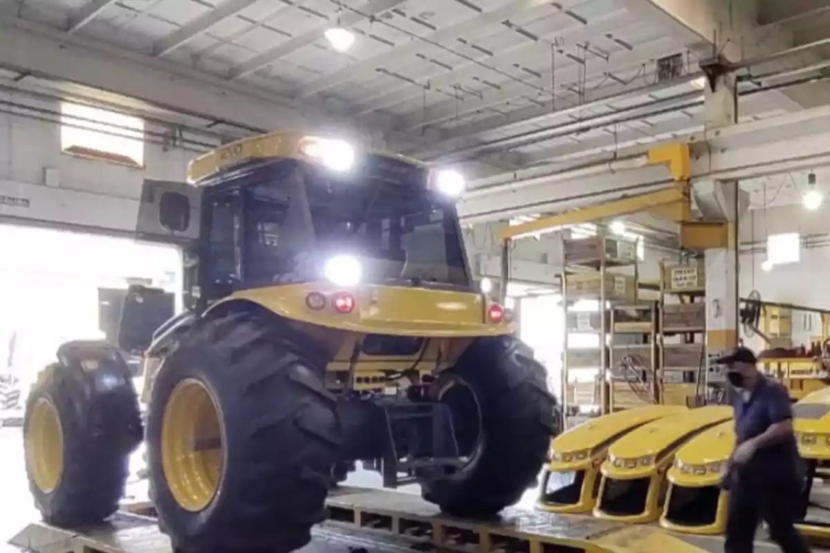 Tractor amarillo de gran tamaño dentro de una fábrica con varias piezas amarillas alineadas y una persona trabajando cerca