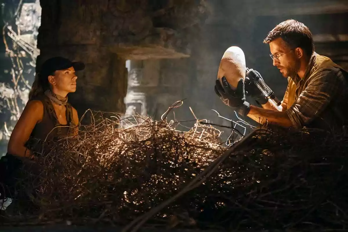 Two people in a cave observe a large, oval object that one of them is holding with gloves, surrounded by branches and in dim light. Two people in a cave observe a large, oval object that one of them is holding with gloves, surrounded by branches and in dim light.