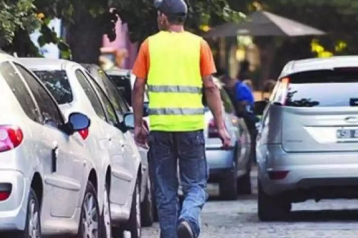 Hombre con chaleco amarillo caminando entre autos estacionados en una calle arbolada Hombre con chaleco amarillo caminando entre autos estacionados en una calle arbolada