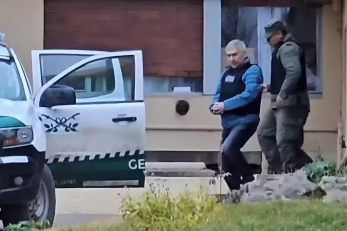Un hombre esposado escoltado por un agente de seguridad junto a una camioneta oficial con la puerta abierta frente a un edificio.