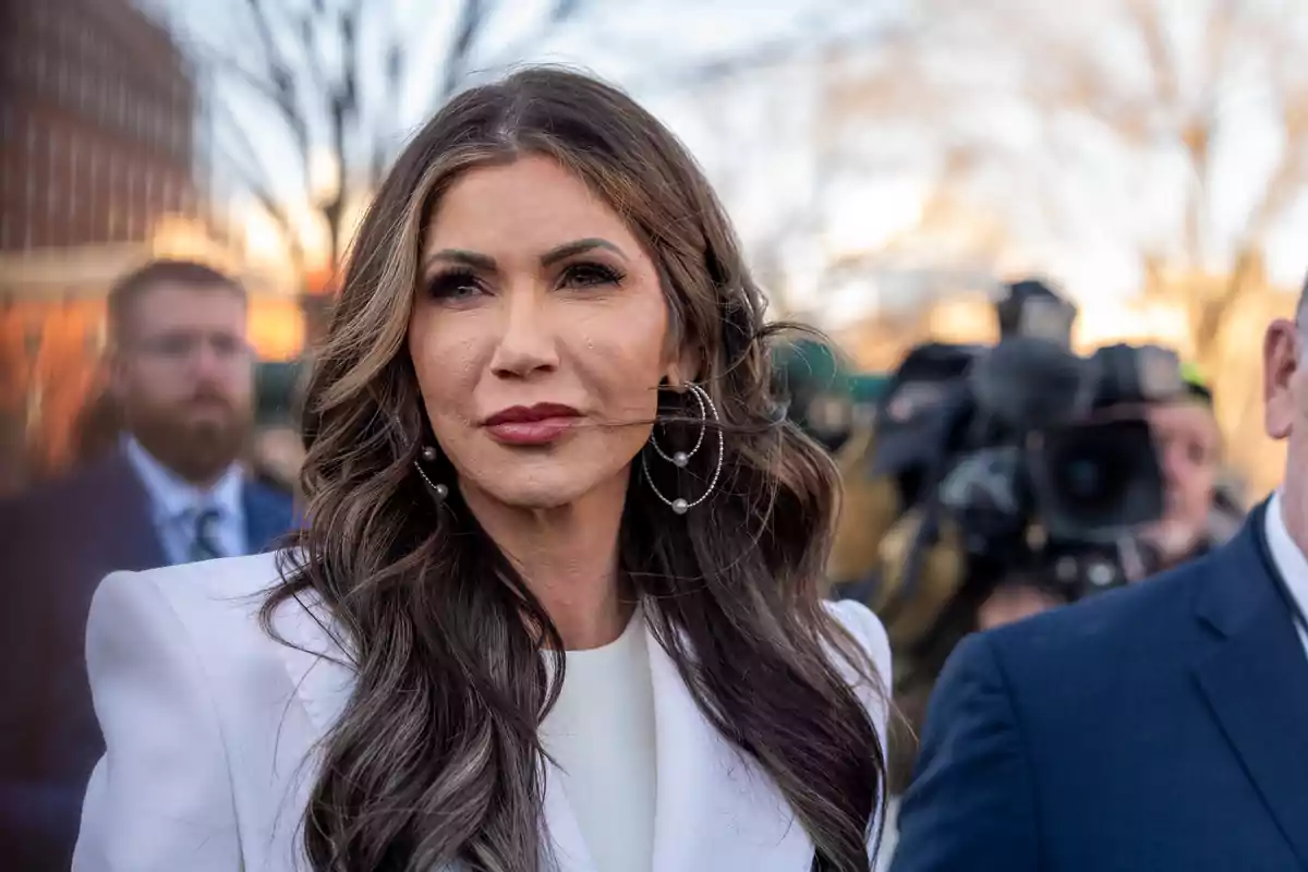 Long-haired, wavy-haired woman with a white jacket and large earrings, surrounded by people and cameras in an outdoor setting