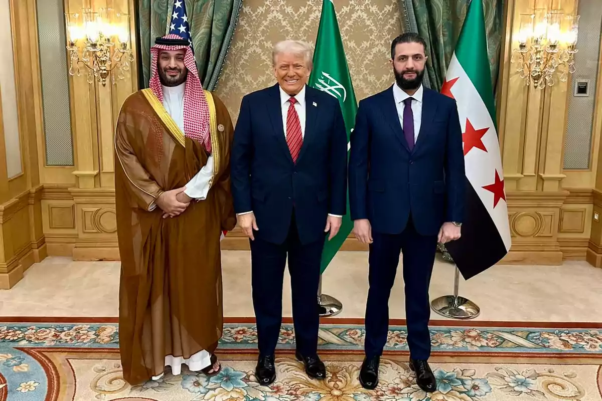 Tres hombres posan juntos en una sala decorada con alfombras y cortinas elegantes, con banderas de Arabia Saudita y Siria de fondo.