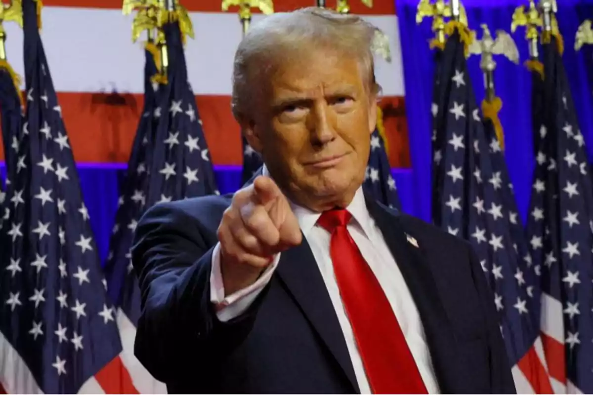 A man in a dark suit and red tie points forward with American flags in the background.