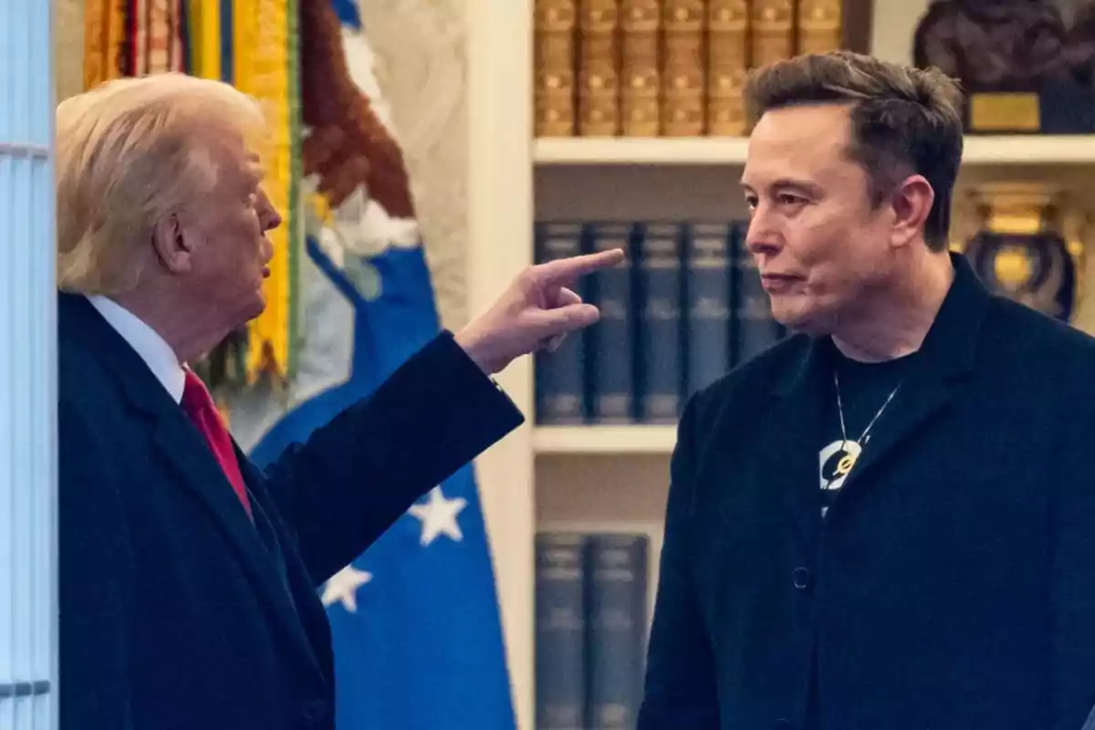 A blond-haired man points his finger at another man with short hair in a room with bookshelves and flags in the background. A blond-haired man points his finger at another man with short hair in a room with bookshelves and flags in the background.