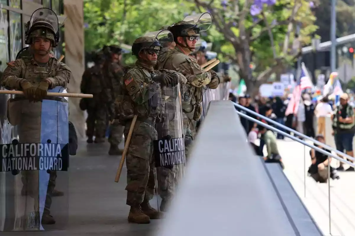 Miembros de la Guardia Nacional de California con equipo antidisturbios y escudos observan una manifestación en la calle mientras personas con pancartas y banderas se reúnen al fondo.