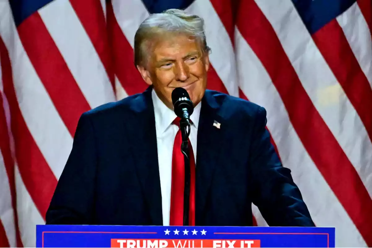 A man in a dark suit and red tie speaks in front of a microphone with American flags in the background.