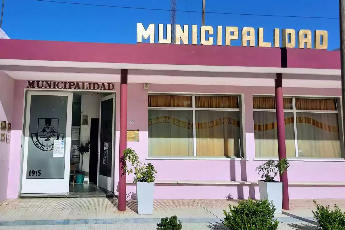 Fachada de un edificio municipal pintado de rosa y blanco con letras doradas en el techo que dicen "Municipalidad" y una bandera ondeando a la izquierda.