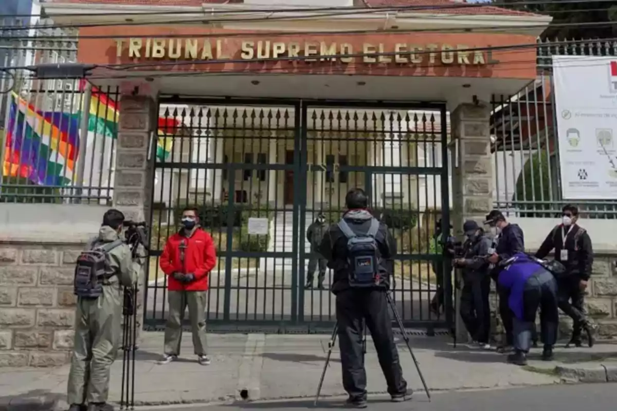 Un grupo de periodistas y camarógrafos se encuentra frente a la entrada del Tribunal Supremo Electoral, con banderas coloridas ondeando a un lado y un cartel informativo visible en la pared.