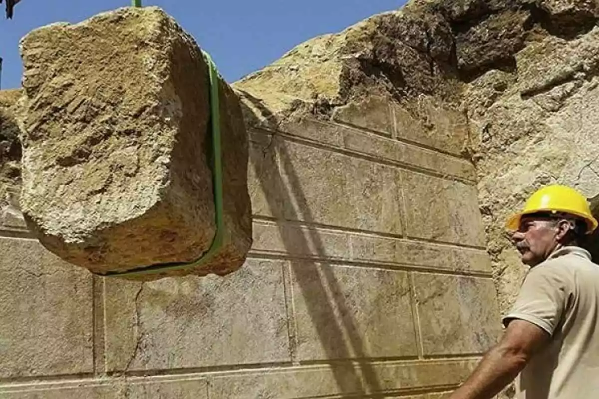 A worker with a helmet watches a stone block suspended by a strap at a construction site. A worker with a helmet watches a stone block suspended by a strap at a construction site.