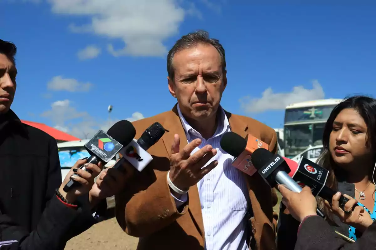 Un hombre hablando con varios micrófonos de diferentes medios de comunicación a su alrededor en un entorno al aire libre.